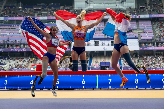Three people with lower limb prosthetics jumping up and down with flags in a stadium.