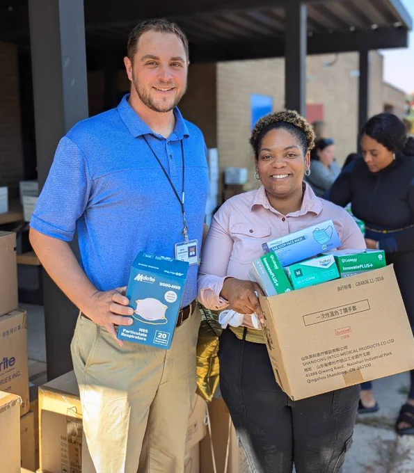  Two people standing outdoors holding boxes of supplies, including masks. They are surrounded by additional boxes, indicating a distribution or donation event.