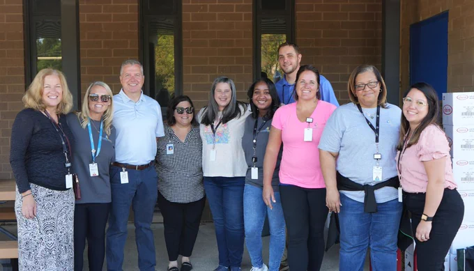A group of ten people, some wearing badges, are standing together outdoors in front of a building and smiling at the camera.