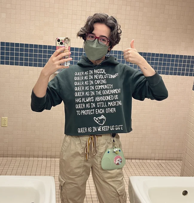 Bathroom selfie at work, wearing khaki cargo pants, a green kn95, and a short green hoodie. The hoodie has white screen printing writing that says “Queer as in radical
Queer as in revolutionary
Queer as in caring
Queer as in community 
Queer as in the government has always abandoned us
Queer as in still masking to protect each other
Queer as in we keep us safe”, with a simple illustration of a mask between the last two lines.