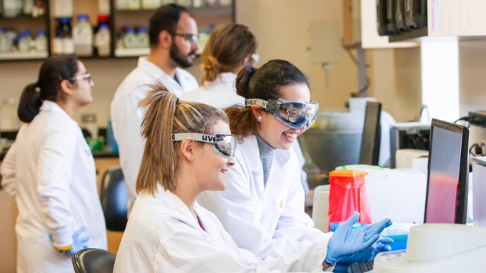 In a classroom laboratory, two students in lab coats, goggles and gloves watch a computer screen, smiling together. Three other students are in the background.