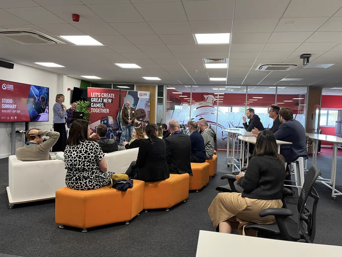 A group of people in a studio environment gathered around as a few people present and talk to the group.