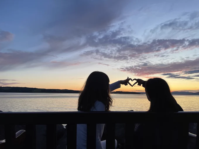 The sun is setting in Largs. Sitting by the sea on a bench are two young girls, part of an Ellen MacArthur Cancer Trust sailing adventure, and with their hands they are creating the outline of a heart on the twilight sky. 