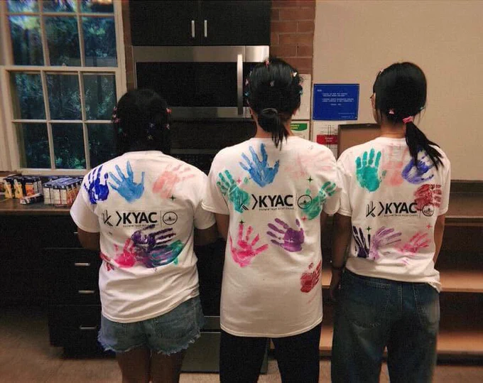 Three youth wearing matching white t-shirts facing away from the camera. On the t-shirts are multicoloured hand prints made out of paint, plus the letters "KYAC."