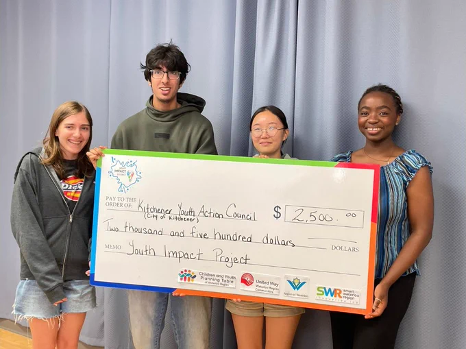 A group of four young people smiling and holding a giant cardboard cheque. The cheque is written out to "Kitchener Youth Action Council" from the Youth Impact project.