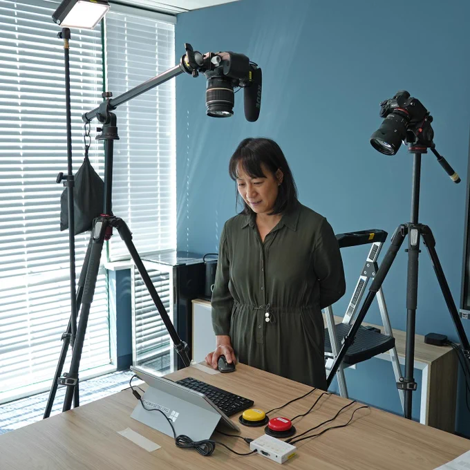 Pearl stands at a desk with a tablet hooked up to a keyboard, and a switch interface with 2 switches. A mounted camera points down towards the tablet screen.  And another is mounted behind her looking over her shoulder.