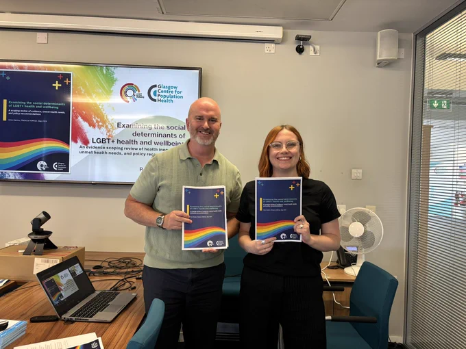 Chris and Rebecca stand in front of a presentation slide which reads “examining the social determinants of LGBT  health and wellbeing. They are both smiling at the camera, and holding copies of the recently published scoping review produced by GCPH and LGBT Health and Wellbeing.