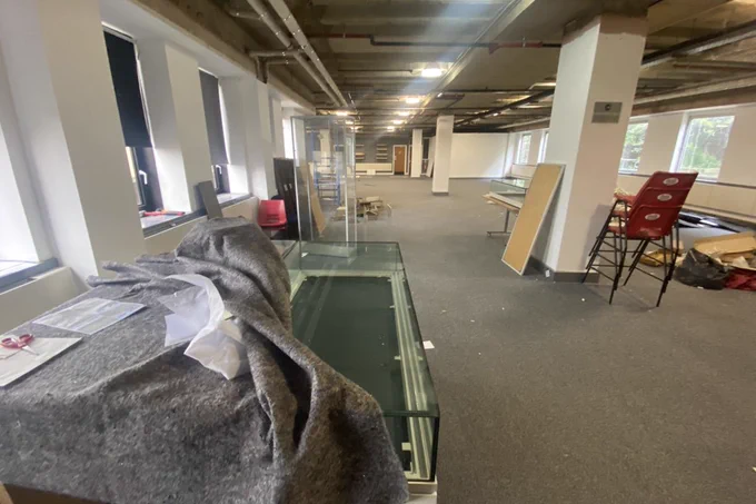 Looking across a large and mainly empty room. Empty display cabinets are on the left and right, one covered in a balnket, there is a stack of red plastic chairs on the right and empty cardboard boxes and other items are on the floor.