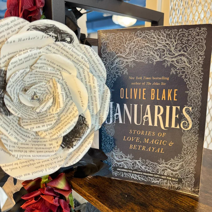 A copy of Januaries by Olivie Blake standing on a shelf next to a rose made of the pages of a book.