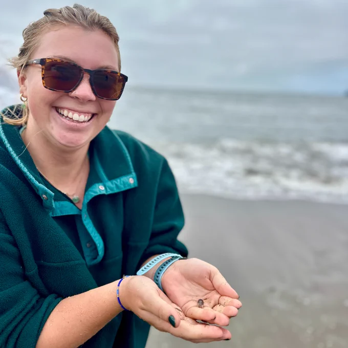 A NatureBridge educator smiling and holding a small crab in their hands with the beach and ocean in the background