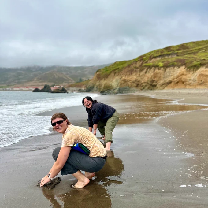 Two NatureBridge educators smiling and bending over the sand near the ocean's edge with bluffs and clouds in the background