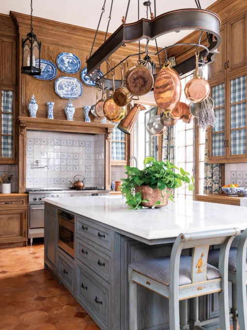 First: A cozy and warm kitchen with wooden cabinets, a pot rack holding copper pots, blue-and-white transferware plates hanging on the wall, gingham curtains in the cabinets, and delft tile on the backsplash.
