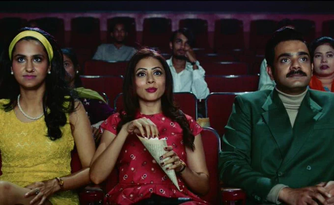 a still from THE QUEEN OF MY DREAMS: 3 people sitting in the cinema, watching the screen, the woman in the middle eats popcorn. Other figures can be seen in the rows behind.  