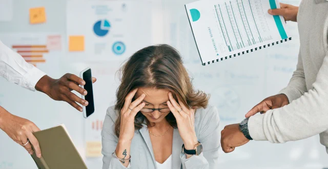 donna al lavoro con le mani sulla fronte in evidente stato di disagio, mentre diverse persone la circondano tenendo in mano telefoni, agende e documenti - Burnout
