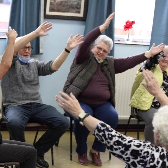 Large group sitting in a circle with their arms in the air, stretching to one side. 