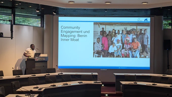 Photo showing a lecture hall. A man is standing at the podium, a large screen shows a slide with a group photo and the title "Community Engagement and Mapping: Benin Inner Moat".