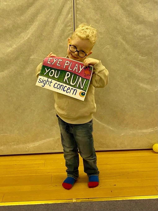 A young boy with bright blond hair and glasses, smiling, holding a sign that says "Eye Play. You Run. Sight Concern."