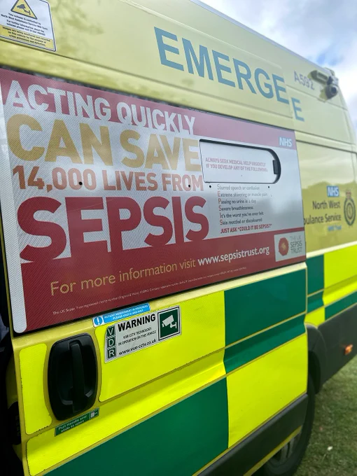 Close-up of a bright, yellow NHS ambulance with a sign highlighting the importance of acting quickly to save lives from sepsis, directing to visit sepsistrust.org for more information.