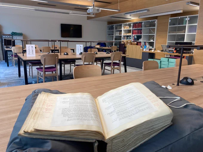 Photo of the Archives Reading Room with an old hardbound book open on a book rest in the foreground. In the background there are chairs, tables with overhead lamps, and bookshelves.