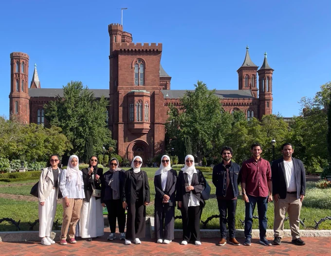 Group of nine people standing in front of the Smithsonian Institution Building, also known as the Castle, on a sunny day.