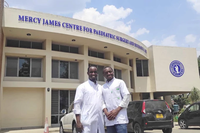 Two men, Alliance Niyukuri, M D, and Bienvenu Mbida, M D, stand smiling in white coats in front of the pediatric surgery center where they trained at Mercy James Centre for Paediatric Surgery and Intensive Care in Blantyre, Malawi.