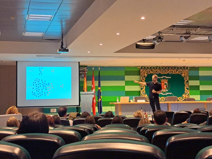 En la imagen se ve a Héctor Ruiz (ropa oscura) con una mesa de conferencias al fondo. En tercer plano hay una pared con el escudo de la Universidad Autónoma de Madrid de fondo, ubicado en una pared pintada a franjas rectangulares de distintos tonos de verde.
A su derecha aparece una pantalla con una proyección que ilustra un modelo de redes de memoria .
En primer plano se ve el patio de butacas con algunos ocupantes en ellas.
