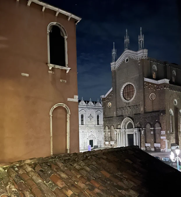 Venice at night. A photo of a tiled roof made of aged brown tiles at the bottom of the picture with what looks to be a church in a square or some such building in the main right side and an orange brown wall and white rimmed windows in that wall.