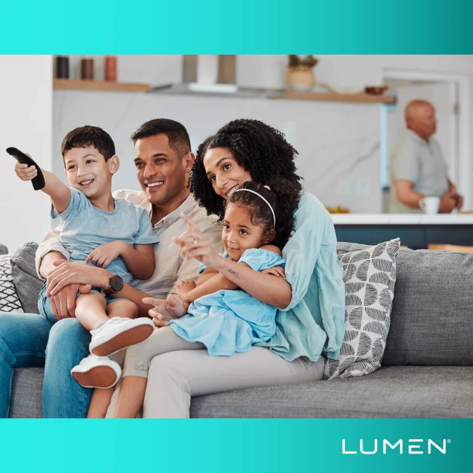 A family of four sitting on a couch, smiling and watching TV together. A child points at the screen. In the background, another adult is in the kitchen. The image includes the logo "LUMEN" in the bottom right corner.