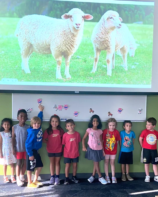 Image of nine small children posing in front of a projector screen showing an image of two sheep.