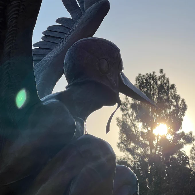 Winged human-like creature with a bird mask perched at sunrise.