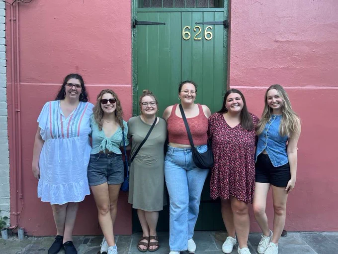 6 women pose in front of a building with pink walls and a green door