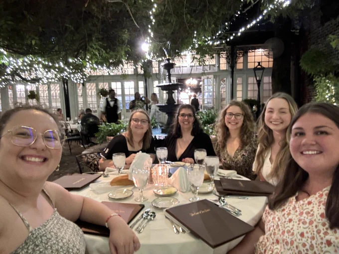 6 women sit around a table outside
 There are vines adorned with white lights above them.