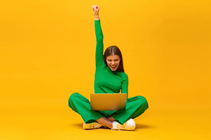 A happy and excited looking young woman is sat cross legged on the floor. She is looking at a laptop and has one fist punched into the air.