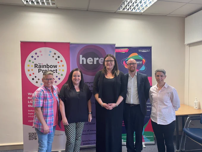Baroness Anderson in the middle of a group picture with 4 other people. Behind them are banners which read The Rainbow Project, here and Cara Friend.
