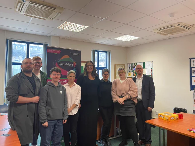 A group shot of 8 people including Baroness Anderson in an office setting. The banner behind them is from Cara Friend.