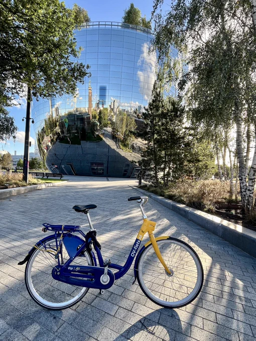 A blue and yellow shared bike stands in a Rotterdam public space lined with trees and plants. A curved and reflective building can be seen in the background.