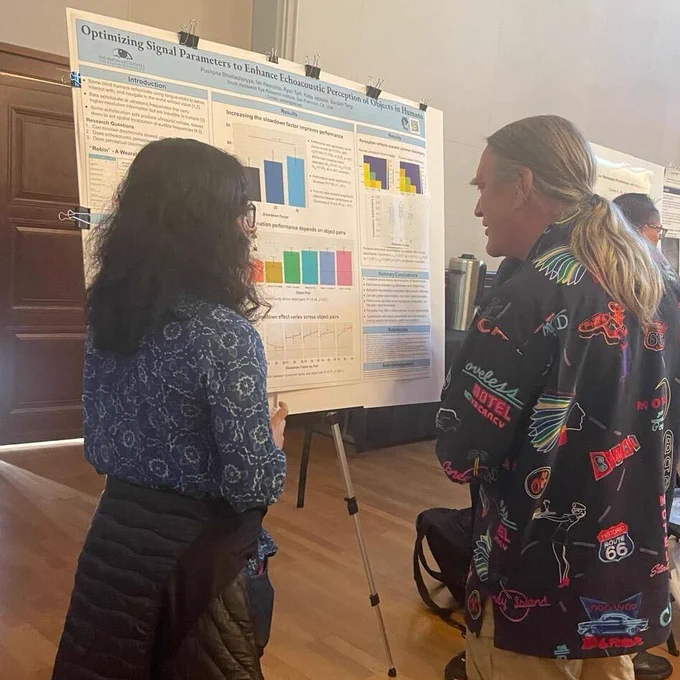 Conference poster titled "Optimizing Signal Parameters to Enhance Echoacoustic Perception of Objects in Humans" being discussed by a long, dark-haired presenter and a visitor with a long gray ponytail and colorful Route-66-themed shirt 