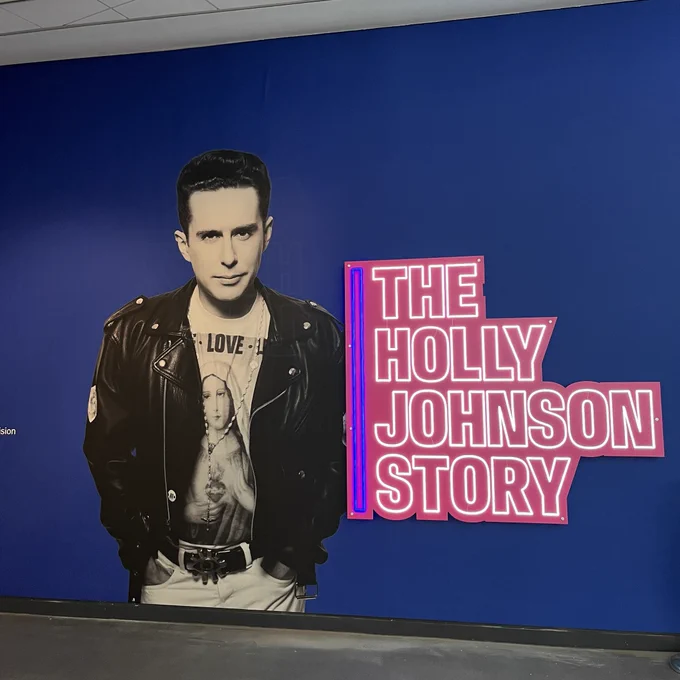 Promotional wall graphic for "The Holly Johnson Story" featuring Holly Johnson in a leather jacket against a blue background with the title in neon pink to the right.