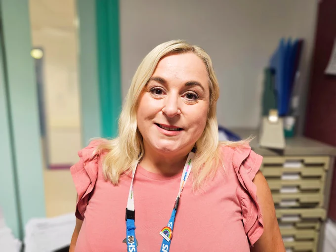 A smiling hospital staff member wearing an ID lanyard stands in a corridor with medical filing cabinets in the background.