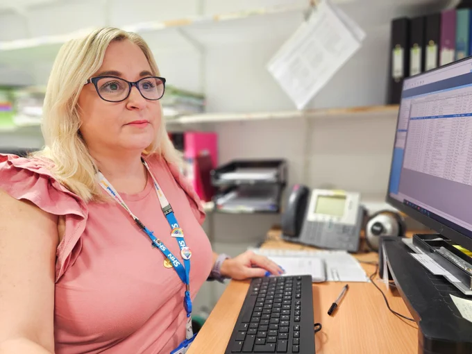 Person with glasses working at a computer in an office, wearing a pink ruffled shirt and an ID badge on a lanyard, surrounded by office equipment and files.