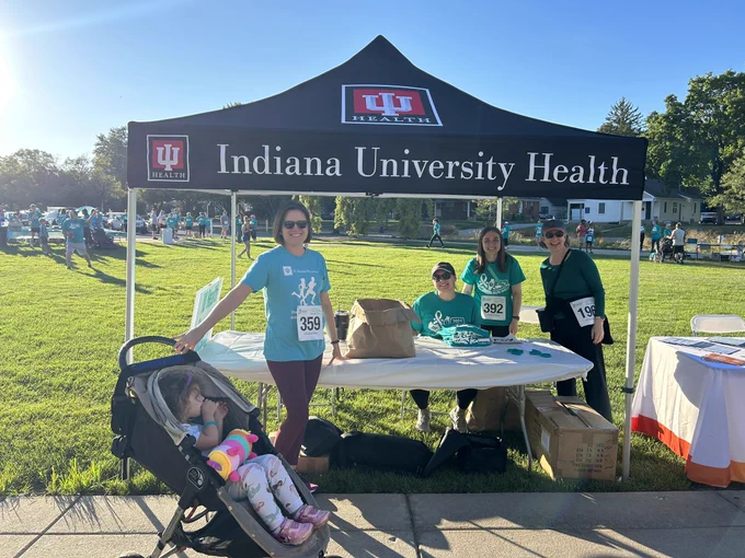 Four women stand and sit at a table under a tent reading Indiana University Health. A child sits in a stroller looking off to the side. Walkers in teal shirts stand in the background on the grass.