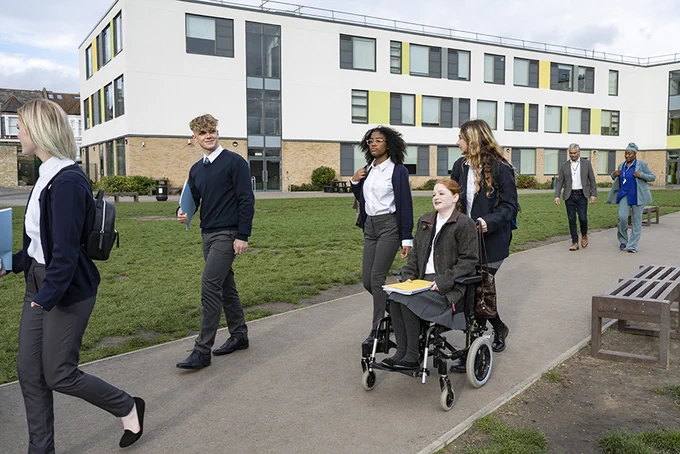 A group of secondary school children walking outside on school grounds. One child is in a wheelchair and is being assisted by a peer.