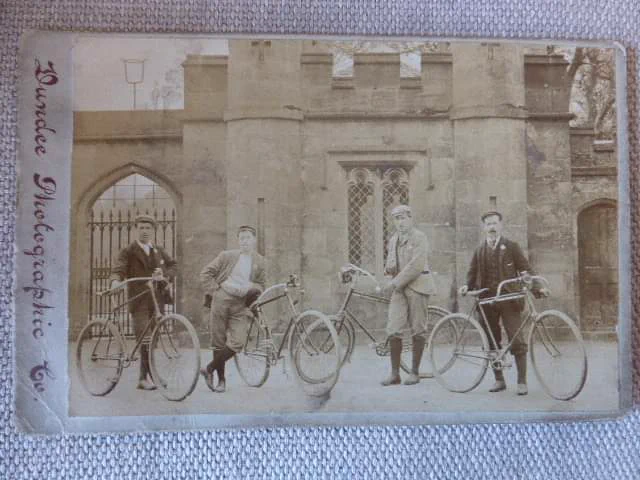 four victorian bicyclists with bikes standing in front a gatebuilding perhaps, with crenulations.