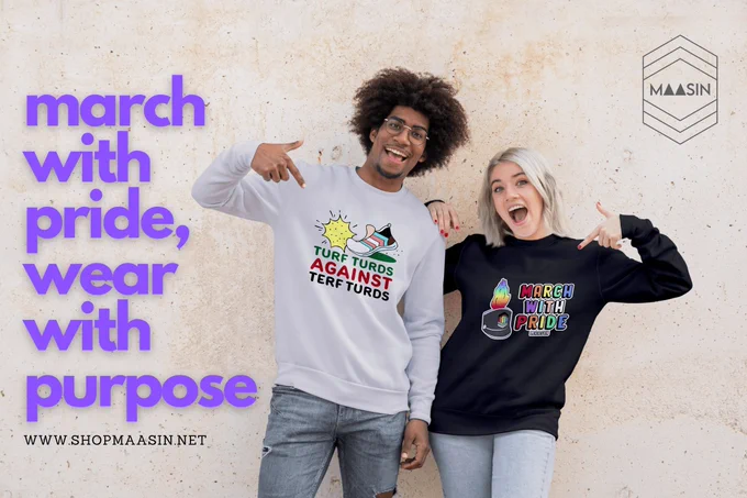 Two individuals stand against a textured wall, smiling and pointing at their sweatshirts. The person on the left wears a light gray sweatshirt with the text 'Turf Turds Against TERF Turds' alongside a graphic of a shoe stepping on a spiked object. The person on the right wears a black sweatshirt with the text 'March with Pride' featuring a colorful, flaming megaphone graphic. The phrase 'march with pride, wear with purpose' is displayed in large purple text on the left side of the image, along with the website URL www.shopmaasin.net. The MAASIN logo is in the top right corner.