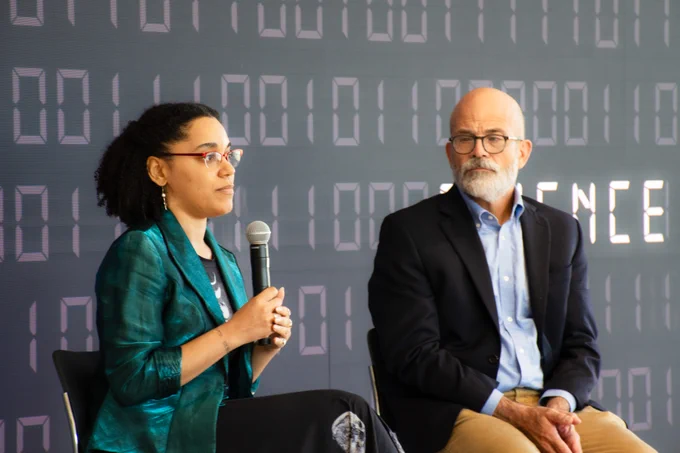 Rafael Alvarado listens to Moiya McTier as she speaks into a microphone, answering student questions. They sit in front of a large navy blue media wall, binary code sifting behind them. 