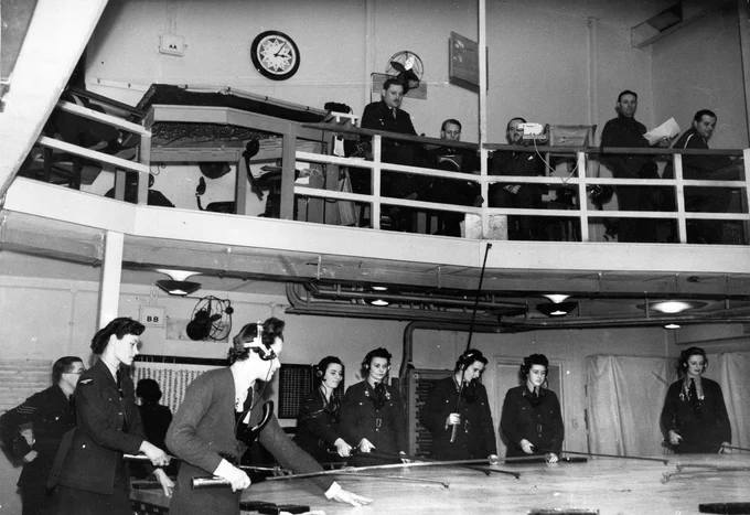 Image shows a black and white photo of plotters working around a large table from a control room.