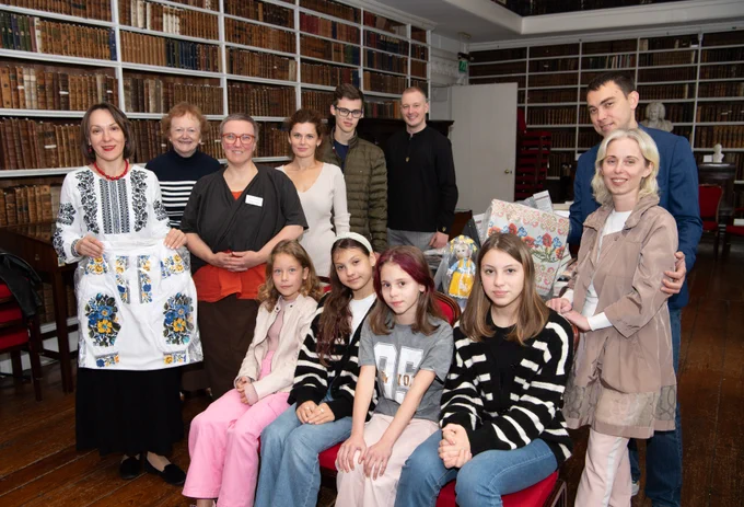 Two staff members and a group of Ukrainian families standing around a table with examples of Ukrainian embroidery in a historic library. | To highlight the 2024 EHOD theme 'Routes, Connections and Networks - Links in our Heritage', local Ukrainian families gave examples of Ukrainian embroidery on loan for display. The families came to visit the display and were met by the Library's Archivist, Thirza Mulder and Assistant Keeper, Carol Conlin.