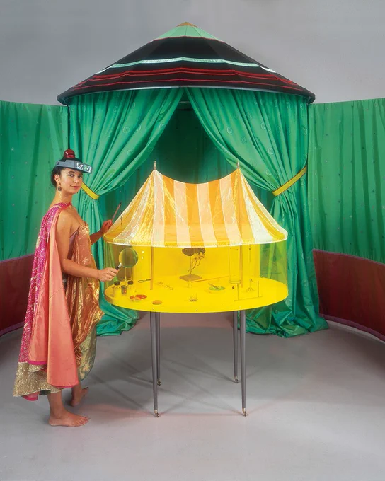 A woman wearing a cape and magnifying headset stands near a large green tent with a smaller yellow arena tent on a table.