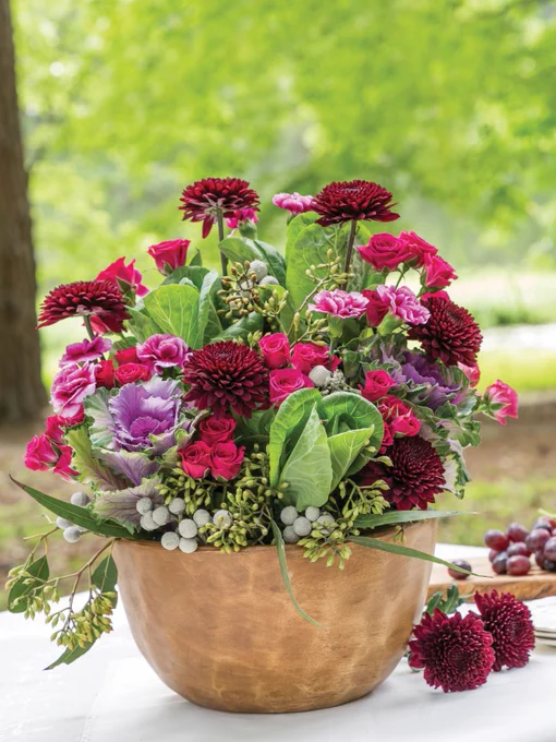 Alt text: A pot holds a purple, burgundy, and fuchsia arrangement of ornamental kale, dahlias, carnations, spray roses, brunia berries, and seeded eucalyptus.
