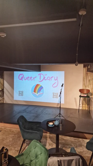 Empty stage.  Microphone on stand. Mini armchairs and tables in front. On wall behind is large screen with 'Queer Diary' written on graph paper.